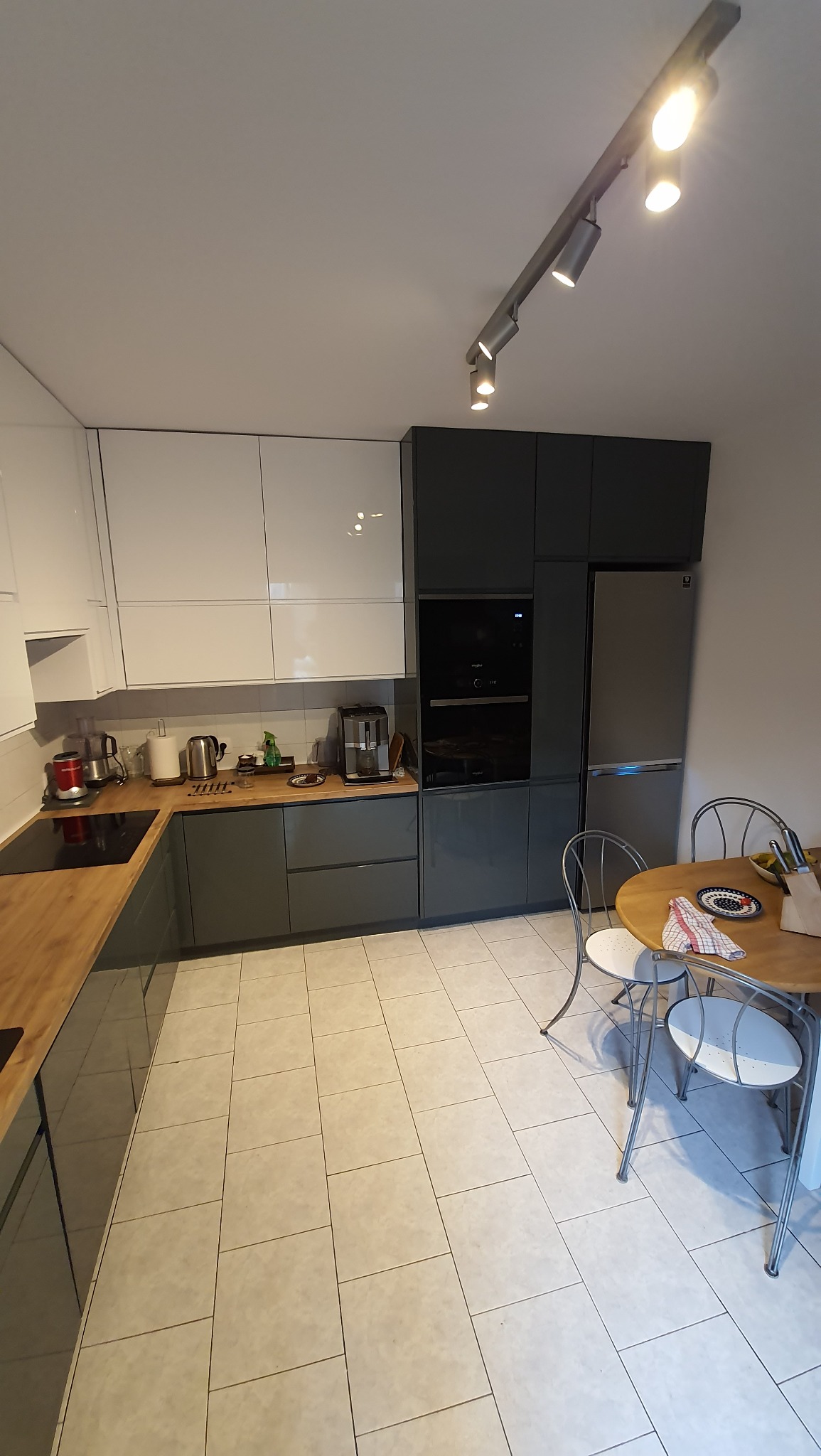 Nowoczesna kuchnia z szarymi i białymi szafkami, drewnianym blatem, okrągłym stołem i metalowymi krzesłami. Oświetlenie sufitowe punktowe.