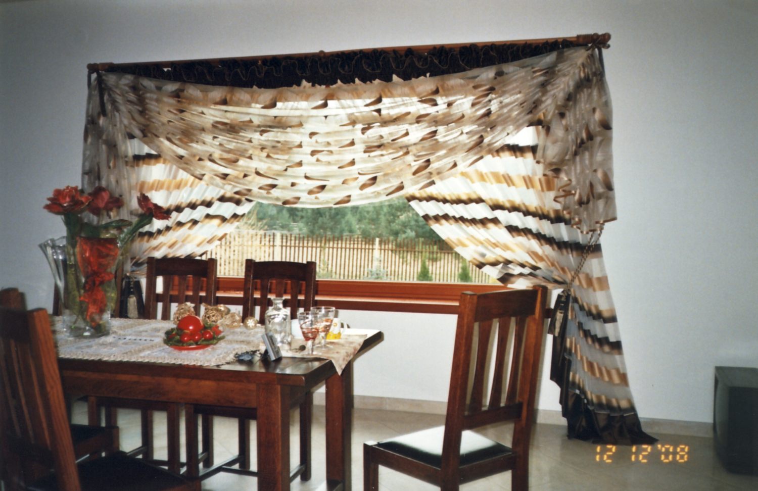 Jadalnia z drewnianym stołem i krzesłami, nakryta do uroczystego posiłku, z dekoracyjnymi firanami w oknie i bukietem czerwonych róż w wazonie.