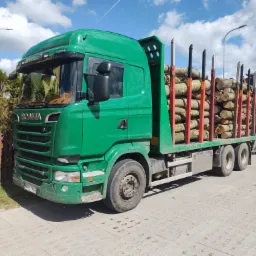 Zielona ciężarówka Scania z ładunkiem drewna na naczepie, zaparkowana w Bożepole Małe, widoczna tablica rejestracyjna.
