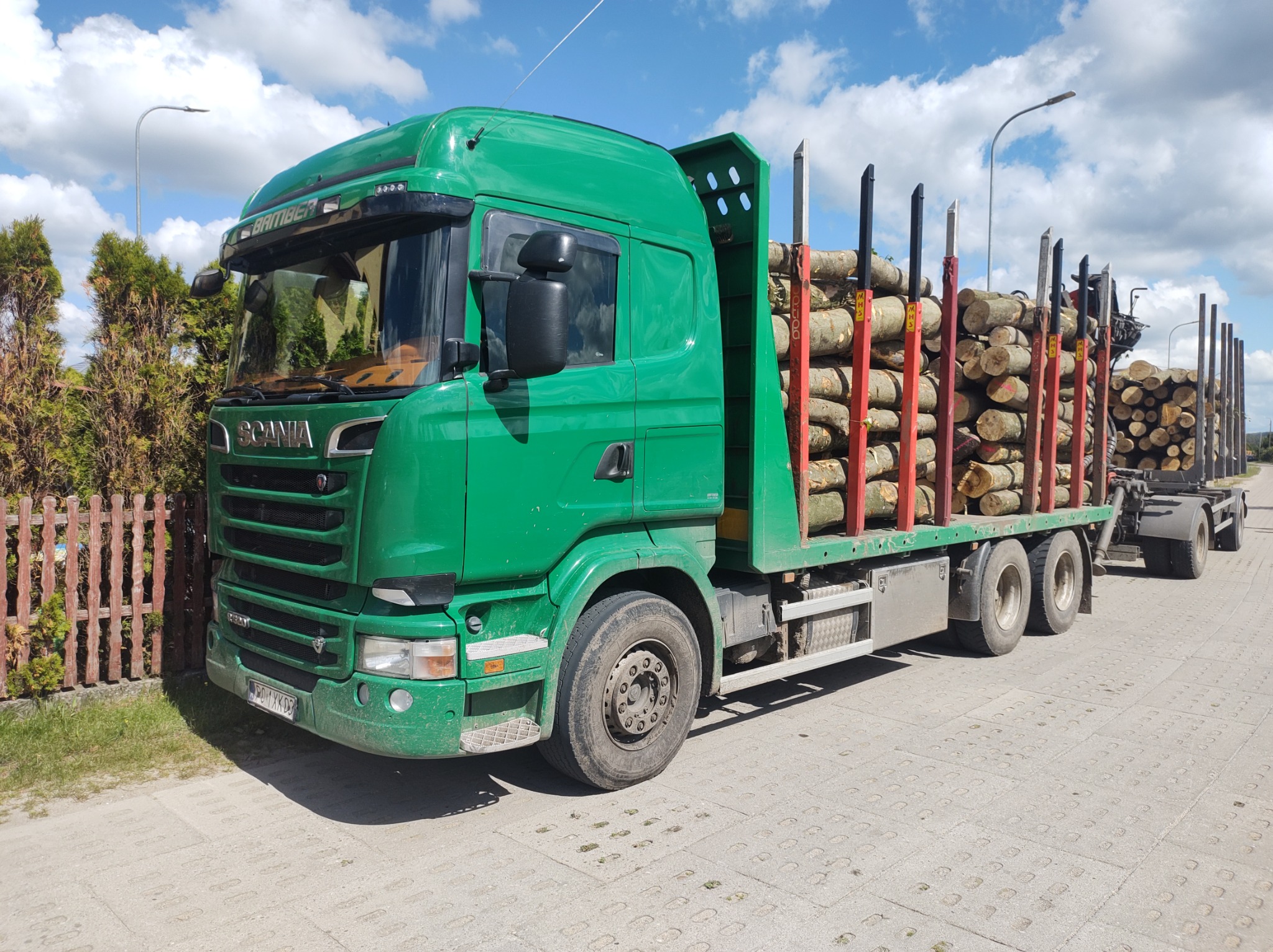 Zielona ciężarówka Scania z ładunkiem drewna na naczepie, zaparkowana w Bożepole Małe, widoczna tablica rejestracyjna.
