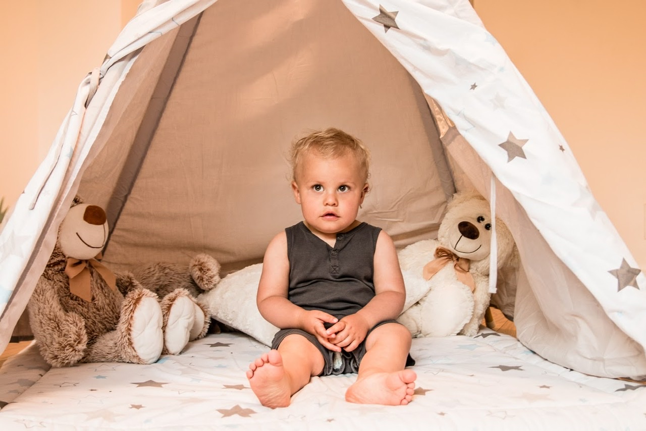 Mały chłopiec siedzi w tipi z pluszowymi misiami po bokach, spoglądając w górę z zaciekawieniem.