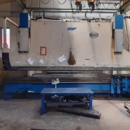 Duża, przemysłowa giętarka do metalu z widocznymi śladami użytkowania, otoczona elementami metalowymi i stołem roboczym w hali produkcyjnej.