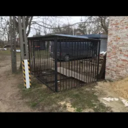 Zewnętrzna, metalowa konstrukcja klatki z prętów, z drewnianą rampą wejściową, na zewnątrz budynku, z widocznym słupem energetycznym obok.