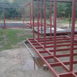 Konstrukcja szkieletowa z profili stalowych pomalowanych na czerwono-brązowy kolor, w trakcie montażu na zewnątrz, widoczne elementy podłogi i zarys przyszłego kształtu budynku.