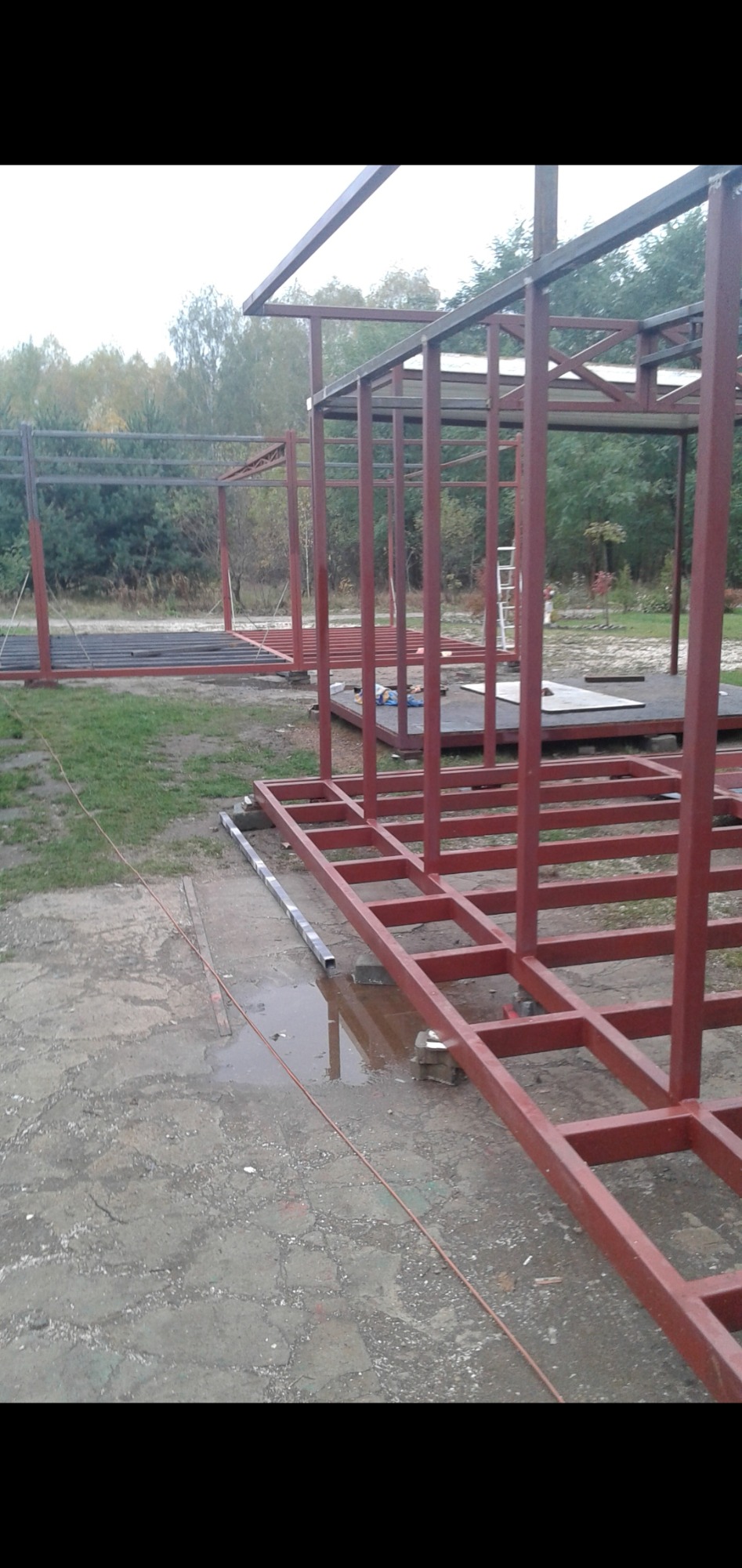 Konstrukcja szkieletowa z profili stalowych pomalowanych na czerwono-brązowy kolor, w trakcie montażu na zewnątrz, widoczne elementy podłogi i zarys przyszłego kształtu budynku.