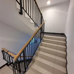 Klatka schodowa w budynku mieszkalnym z jasnymi, wykafelkowanymi schodami, czarną metalową balustradą z drewnianym poręczą i białymi ścianami.