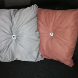 Dwie poduszki dekoracyjne: szara i łososiowa, z ozdobnymi kryształkami pośrodku, ułożone na ciemnoszarym tle sofy.