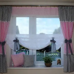 Okno udekorowane w zasłony w różowo-szare kropki z kokardami, roleta rzymska i poduszki w tym samym stylu, na parapecie roślina doniczkowa i ramka ze zdjęciem dziecka.