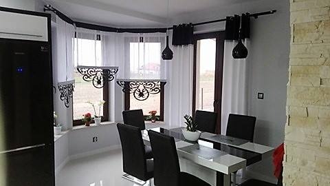 Jadalnia z dużym oknem wykuszowym, udekorowanym białymi, półprzezroczystymi firanami z czarnymi, ażurowymi lambrekinami i czarnymi zasłonami po bokach, wiszące czarne lampy kuliste, nowoczesny stół...