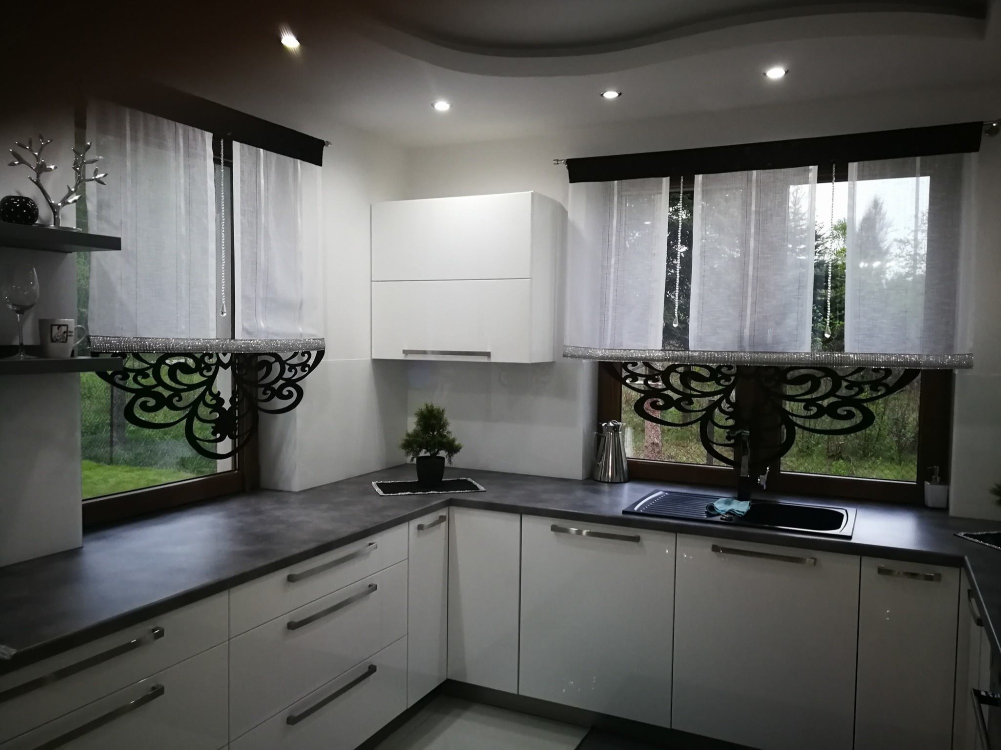 Nowoczesna kuchnia z białymi szafkami i szarym blatem, okna udekorowane białymi zasłonami z czarnymi karniszami i ozdobnymi, ażurowymi panelami dolnymi oraz srebrnymi łańcuszkami.