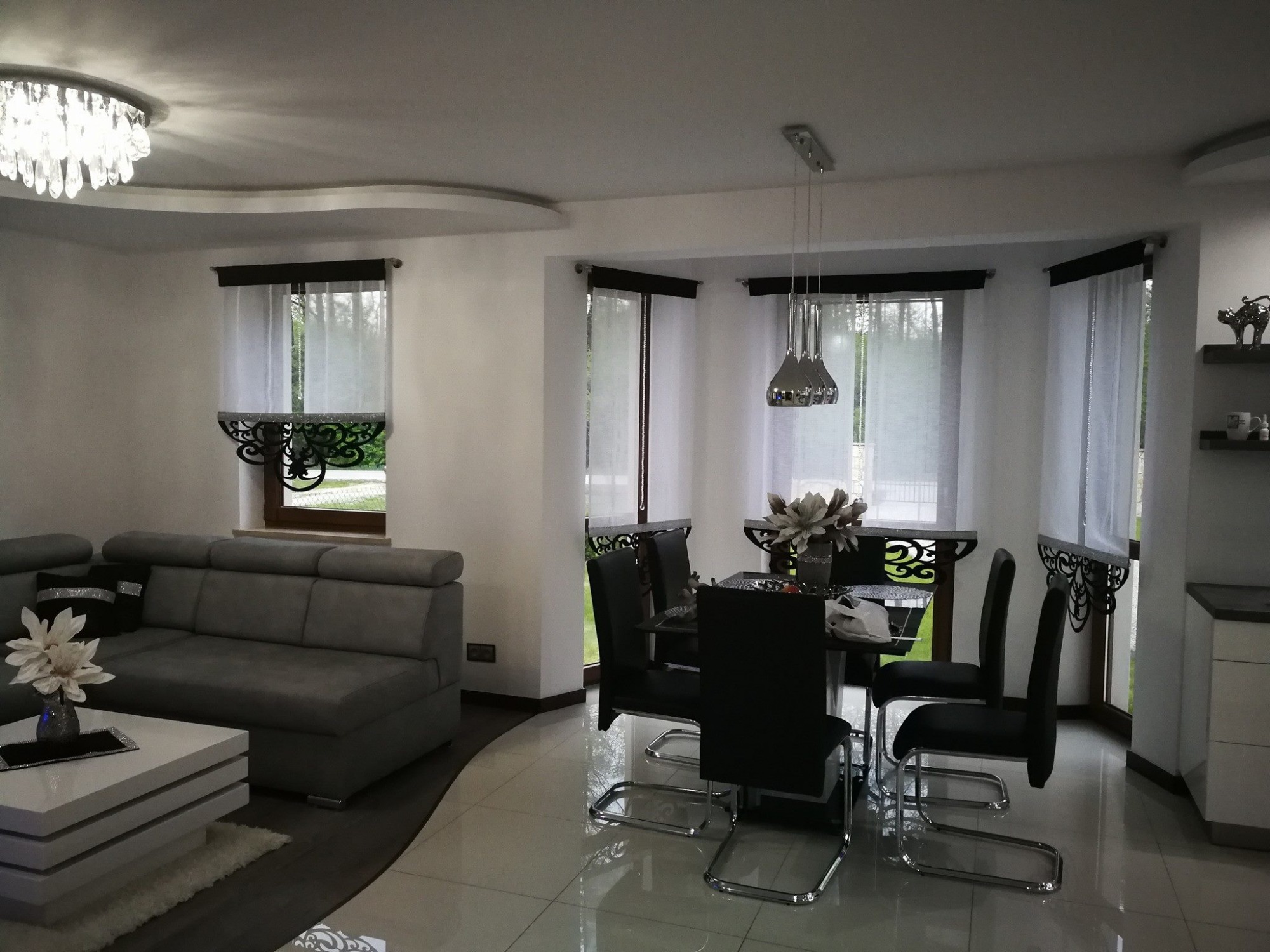 Jasne wnętrze salonu z widokiem na okna z białymi, prześwitującymi zasłonami i ozdobnymi karniszami, szara sofa i biały stolik kawowy z kwiatami, czarne krzesła i stół jadalny z dekoracją kwiatową.