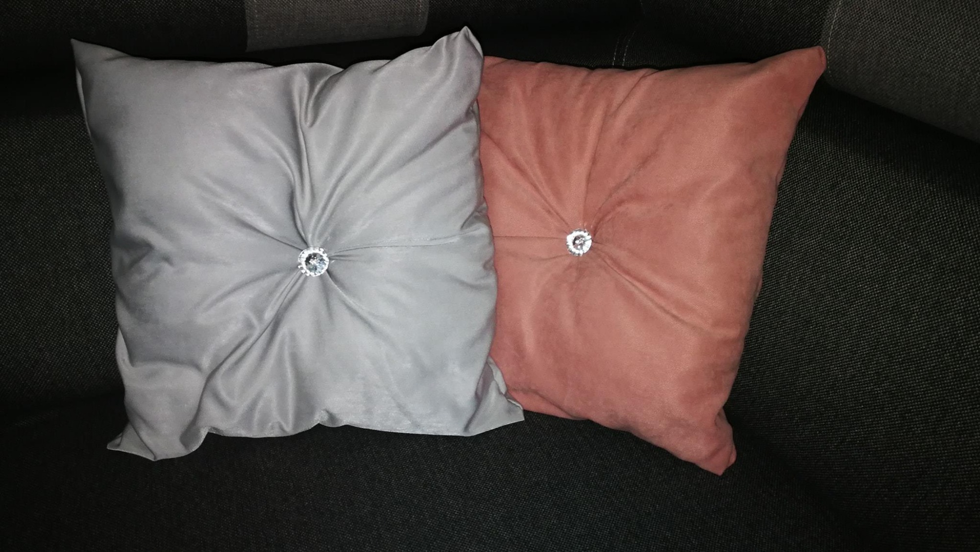 Dwie poduszki dekoracyjne: szara i łososiowa, z ozdobnymi kryształkami pośrodku, ułożone na ciemnoszarym tle sofy.