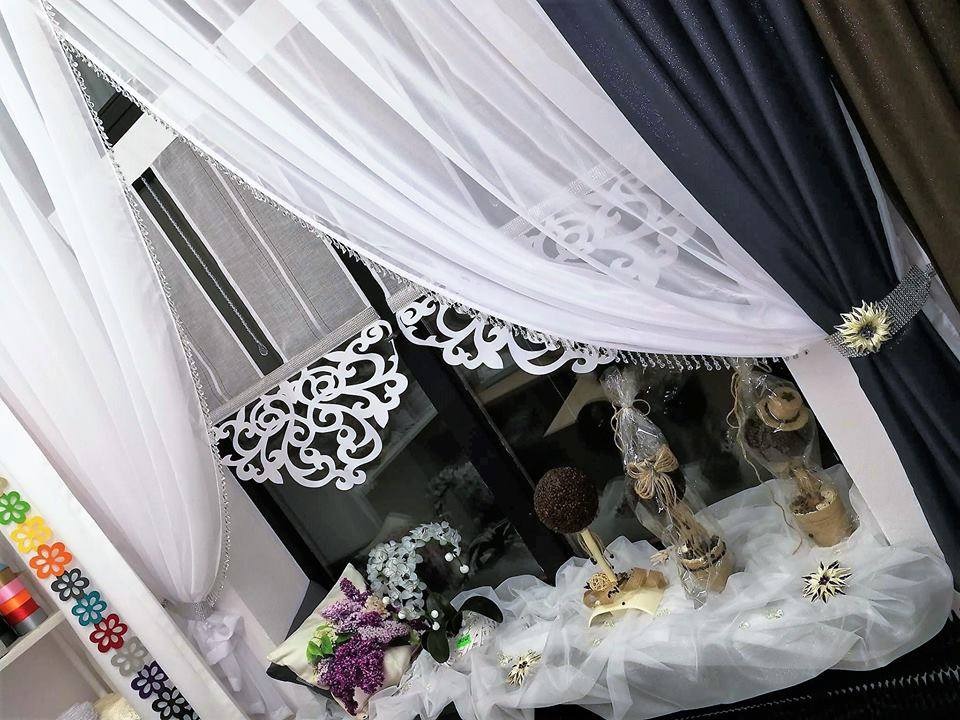 Okno udekorowane białymi, zwiewnymi firanami z ażurowymi wykończeniami i ciemnymi zasłonami spiętymi ozdobnymi klamrami, parapet z dekoracjami florystycznymi i upominkami.