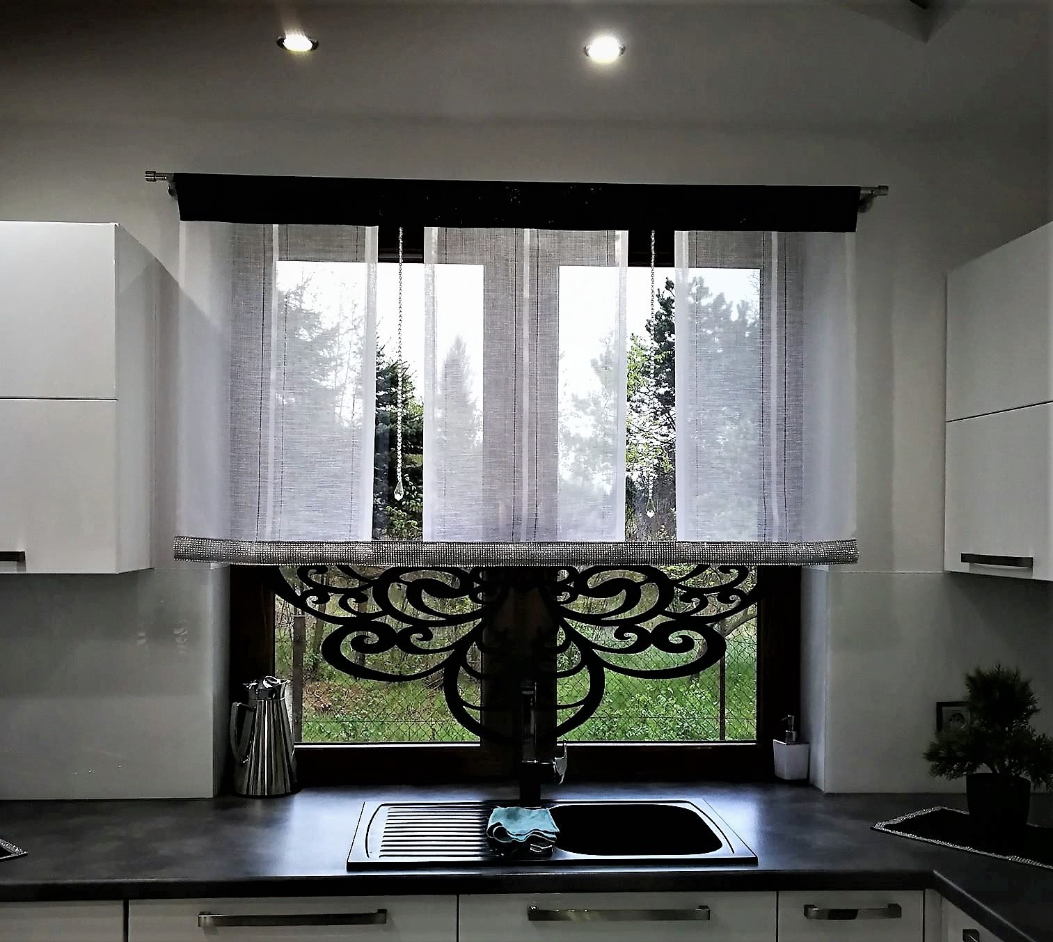 Nowoczesne, białe zasłony z czarnym akcentem i dekoracyjnym, ażurowym panelem dolnym w kuchni z widokiem na zieleń za oknem.