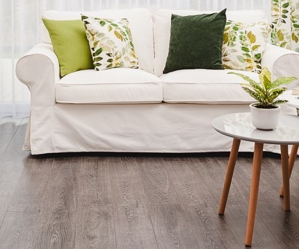 Jasna sofa z zielonymi i liściastymi poduszkami, okryta narzutą, obok okna z zasłoną w liście, stolik kawowy z rośliną w doniczce, na ciemnej podłodze.