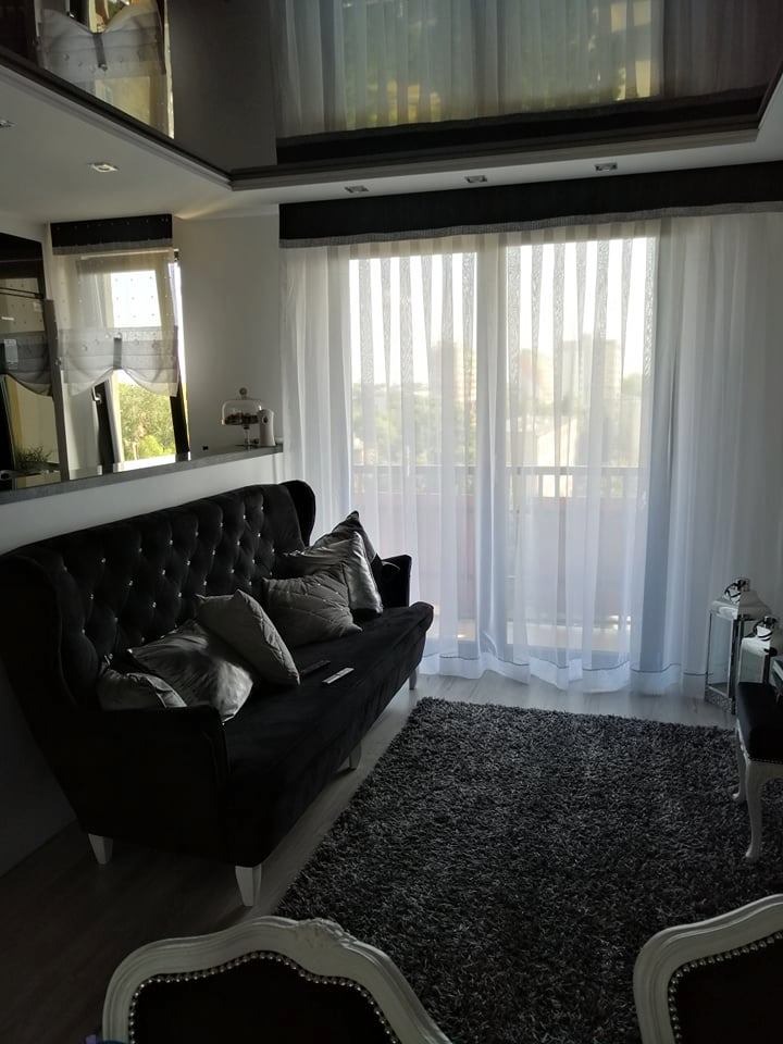 Eleganckie, białe, półprzezroczyste zasłony z czarnym pasem u góry, w jasnym salonie z czarną kanapą w stylu glamour i szarym dywanem.