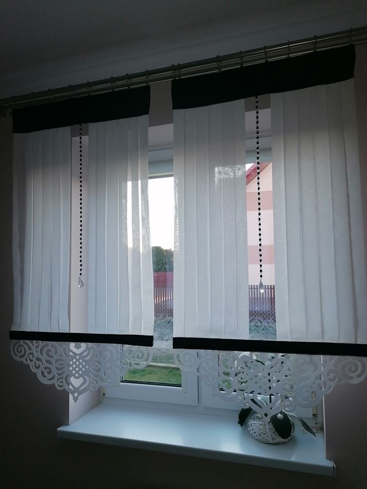 Białe, pionowe żaluzje z czarnymi akcentami i ozdobnym ażurowym wykończeniem dolnej krawędzi, wiszące w oknie. Dekoracyjne łańcuszki z kryształkami. Na parapecie biała doniczka z rośliną.