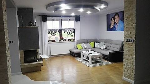 Salon w Radomiu z szarym narożnikiem, białym stolikiem kawowym na wzorzystym dywanie, kominkiem i oknem z białymi zasłonami i dekoracyjnymi lambrekinami.