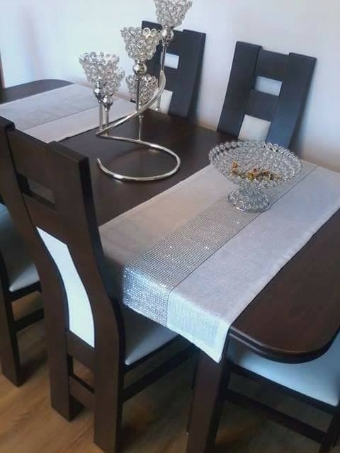 Udekorowany stół z ciemnego drewna z bieżnikiem w odcieniach szarości, zdobionym srebrnymi, błyszczącymi elementami, na którym ustawiono kryształowy świecznik i misę.