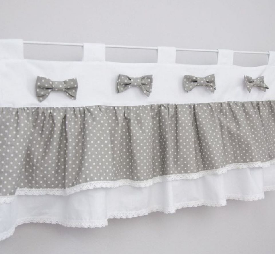 Zasłonka do pokoju dziecięcego z szarej tkaniny w białe kropki, wykończona białą koronką i ozdobiona czterema szarymi kokardkami, zawieszona na białym karniszu.