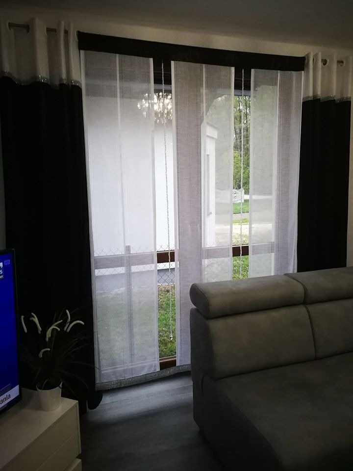 Nowoczesne okno z białymi, transparentnymi panelami i czarnymi zasłonami bocznymi, widoczne przez okno podwórko, w tle kryształowy żyrandol, szara kanapa w kadrze.