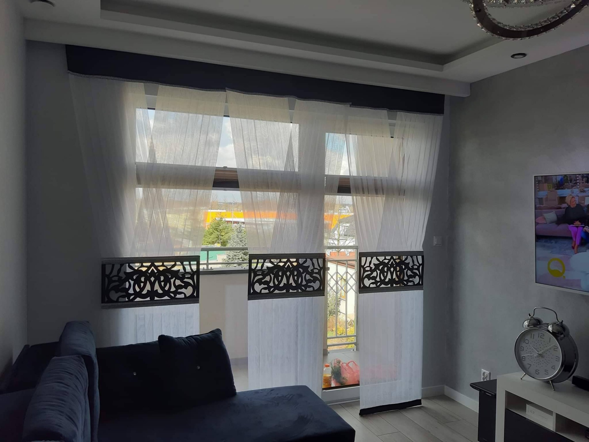 Nowoczesne, białe, półprzezroczyste panele okienne z ażurowymi, czarnymi dekoracjami w górnej i dolnej części, widoczne z wnętrza salonu z ciemną sofą i zegarem na szafce.