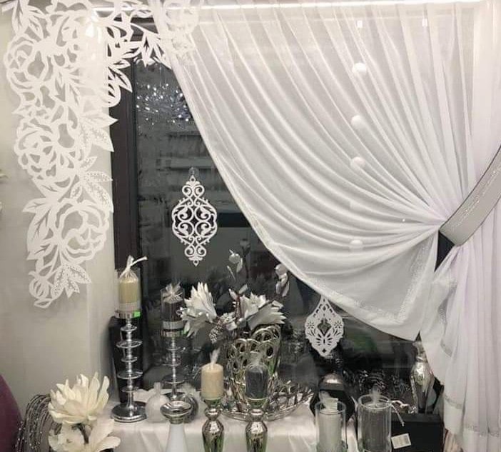 Aranżacja okna z białą, zwiewną firaną i dekoracyjnymi wycinankami na szybie i ścianie, uzupełniona srebrnymi świecznikami i wazonami z kwiatami.
