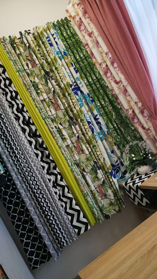 Ekspozycja różnorodnych tkanin na zasłony: geometryczne wzory, motywy roślinne i kwiatowe, gładkie materiały w pastelowych kolorach. Widoczne próbki ułożone pod kątem, prezentujące bogaty wybór...