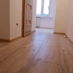 Ułożone panele podłogowe w odcieniach brązu z widoczną teksturą drewna, prowadzące w głąb korytarza z beżowymi ścianami, drewnianą ościeżnicą i drzwiami z przeszkleniami, oraz widocznym fragmentem...