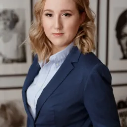 Portret kobiety w niebieskim garniturze i jasnej koszuli, z blond włosami do ramion, na tle ściany z czarno-białymi zdjęciami w ramkach.