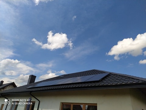 Panele słoneczne na ciemnym dachu domu jednorodzinnego, na tle błękitnego nieba z białymi obłokami.