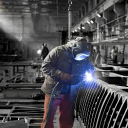 Pracownik w ochronnej masce spawalniczej łączy metalowe elementy łukowej konstrukcji w hali produkcyjnej, iskry i dym w powietrzu.