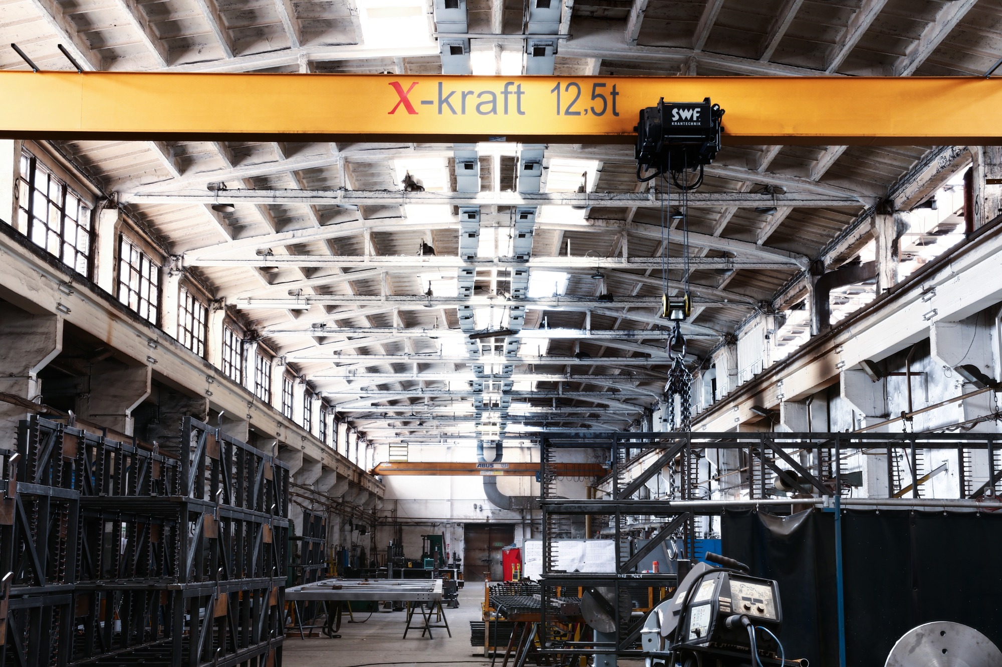Wnętrze hali produkcyjnej z suwnicą bramową X-kraft 12.5t, widoczne konstrukcje metalowe, spawarka i inne urządzenia warsztatowe.