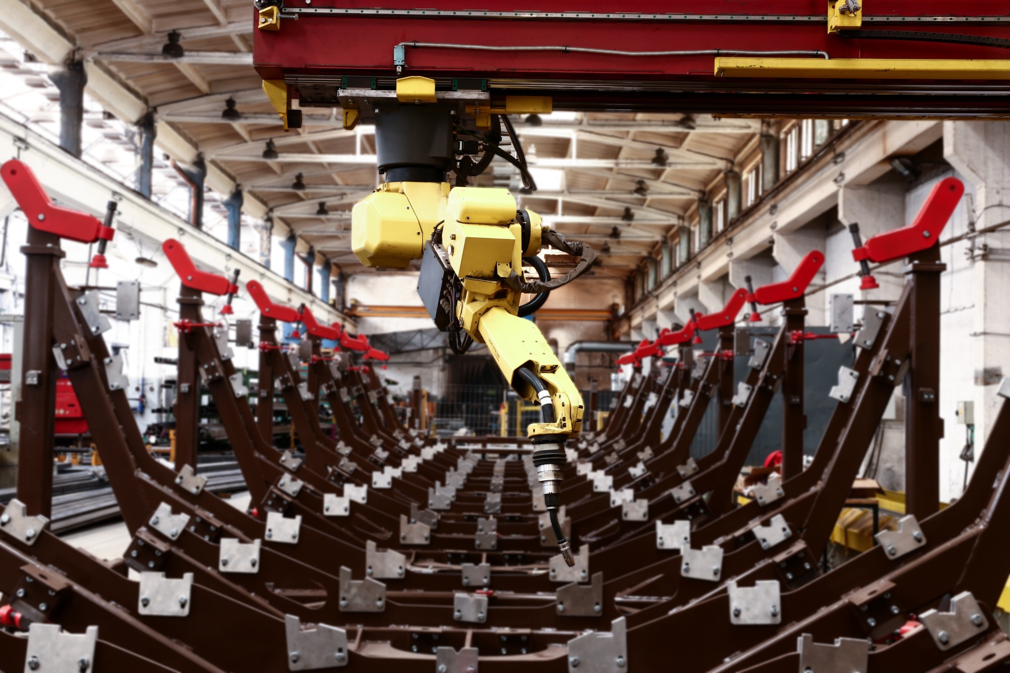 Żółty robot przemysłowy zawieszony nad rzędem brązowych, metalowych konstrukcji z czerwonymi elementami, w hali produkcyjnej.