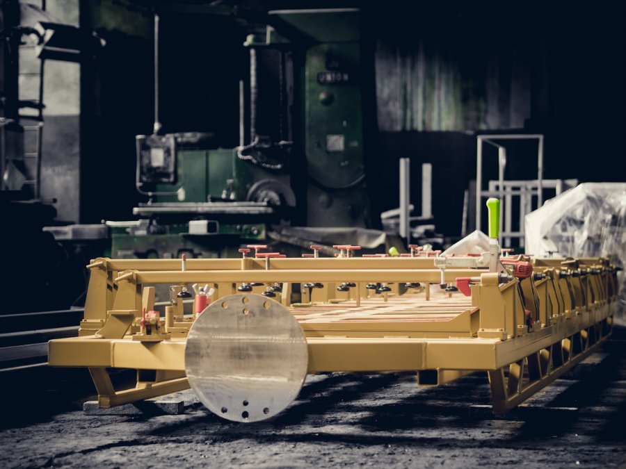 Żółta, metalowa platforma z regulowanymi elementami, okrągłym dyskiem i zielonym uchwytem na tle ciemnego warsztatu z maszynami.