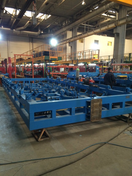 Duża, niebieska rama z licznymi mocowaniami w hali produkcyjnej, widoczny pracownik w tle i instalacje pod sufitem.