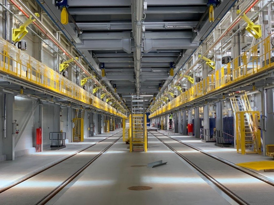 Wnętrze hali produkcyjnej z suwnicami i pomostami roboczymi, widok wzdłuż torowiska, jasne oświetlenie.