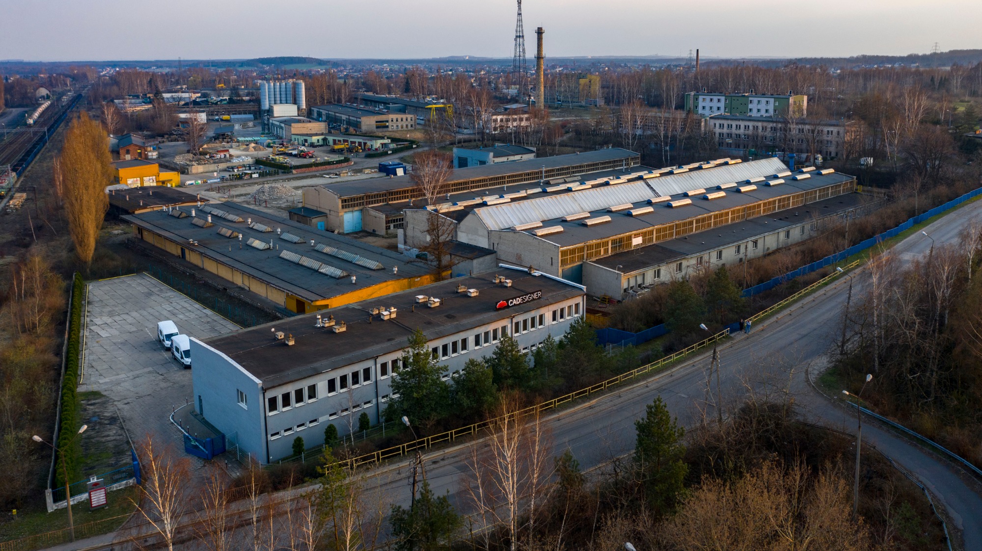 Widok z lotu ptaka na kompleks hal przemysłowych z logo firmy CADesigner na dachu jednego z budynków, z widocznymi elementami infrastruktury kolejowej i drogowej w tle.