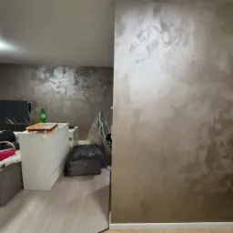 Ściana w salonie z dekoracyjną, metalizowaną farbą w odcieniach beżu i szarości, z widoczną fakturą. W tle widoczny fragment umeblowanego pokoju.