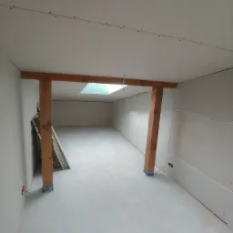 Pomieszczenie w trakcie prac wykończeniowych: surowy stan ścian z płyt kartonowo-gipsowych, drewniana konstrukcja nośna, widoczne łączenia płyt na suficie, przygotowane do dalszych prac.