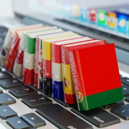 Rząd słowników językowych z flagami narodowymi na grzbietach, ułożonych na klawiaturze laptopa, z widocznym ekranem w tle.