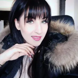 Portret kobiety z ciemnymi włosami i grzywką, ubranej w kurtkę z futrzanym kołnierzem, uśmiechniętej i patrzącej w obiektyw.