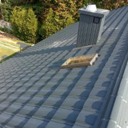 Szary dach z blachy trapezowej, widoczny komin wentylacyjny, okno dachowe, elementy obróbki blacharskiej i przygotowane drewno na zewnątrz.