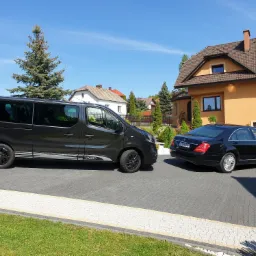 Czarny bus Opel Vivaro z białymi pasami i czarnymi felgami oraz czarna limuzyna Mercedes zaparkowane na posesji przed domem w słoneczny dzień.