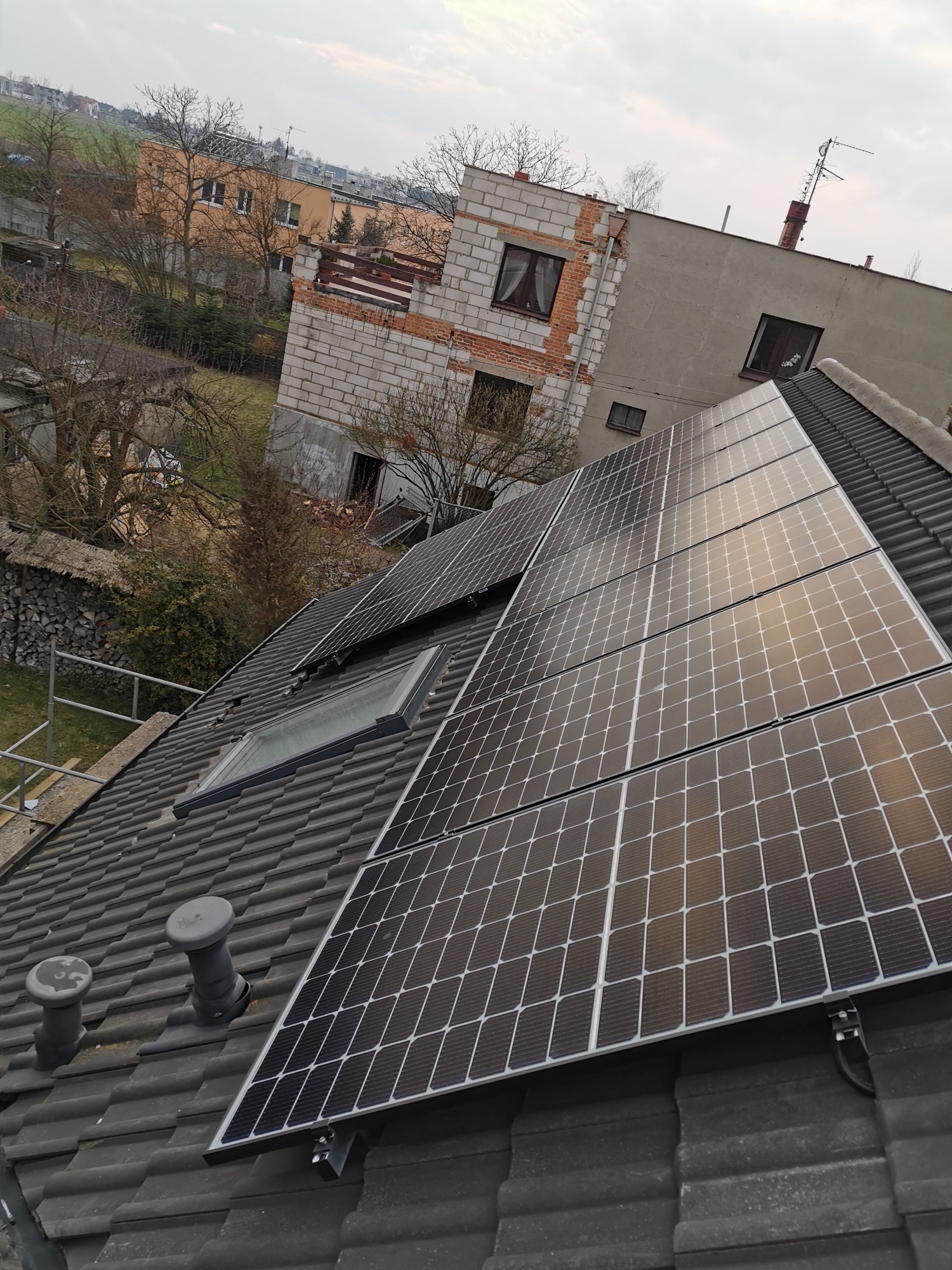 Panele słoneczne na dachu domu z widocznymi kominami wentylacyjnymi i oknem dachowym, w tle budynek w stanie surowym otwartym.