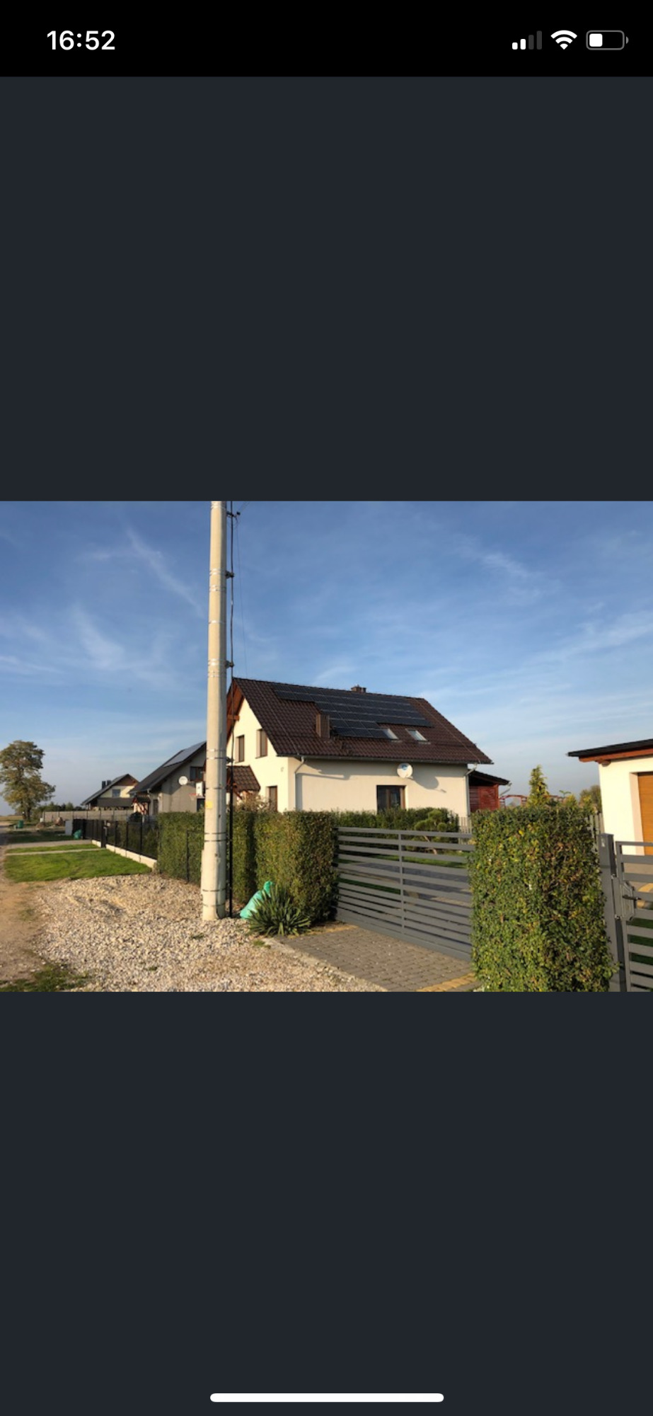 Dom jednorodzinny z brązowym dachem i panelami słonecznymi na dachu, widok z zewnątrz, ogrodzenie z żywopłotu, słup energetyczny na pierwszym planie.