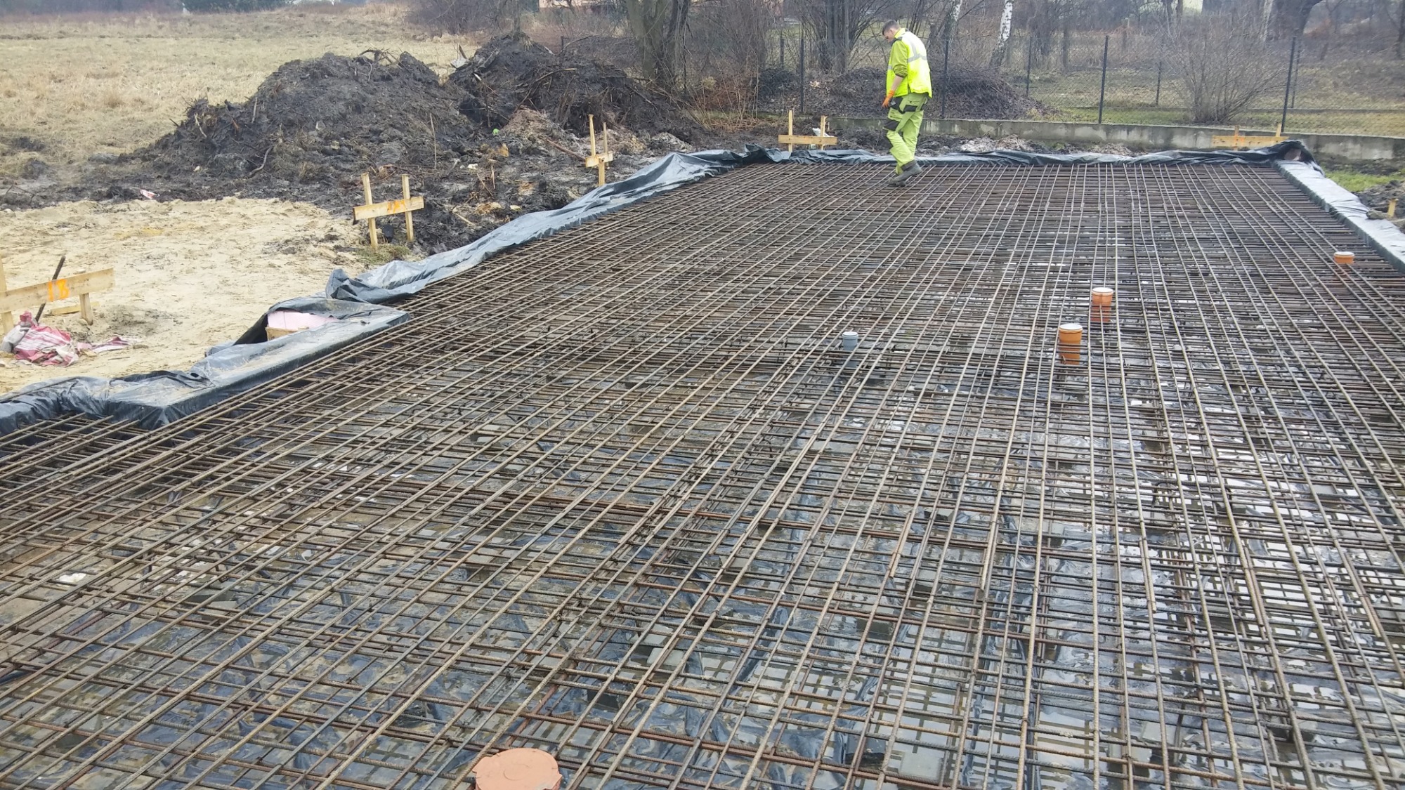 Zbrojenie płyty fundamentowej z wystającymi rurami kanalizacyjnymi, widoczny pracownik w odblaskowej kamizelce i sterta ziemi w tle.
