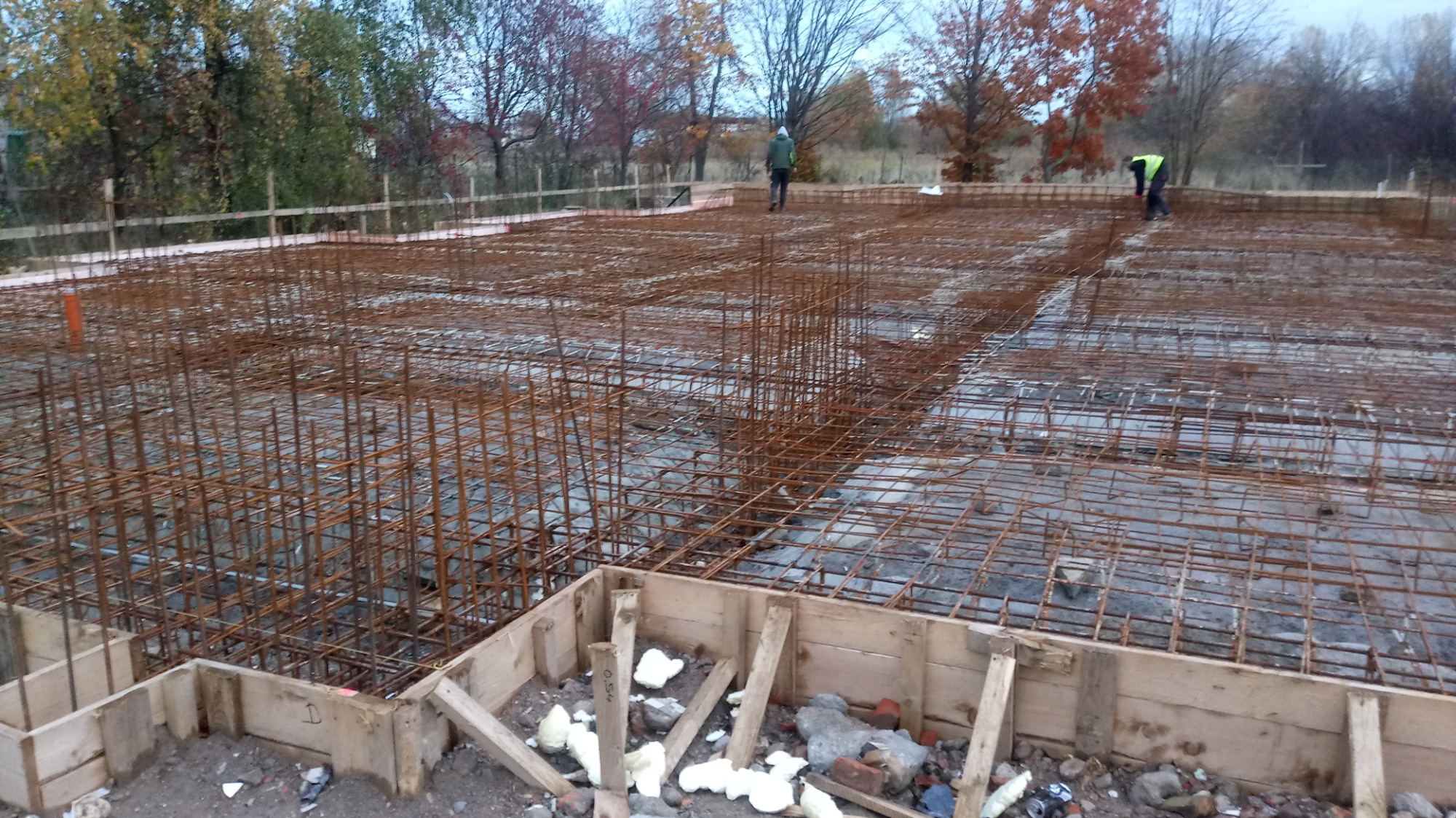 Zbrojenie płyty fundamentowej z widocznym szalunkiem drewnianym i dwoma pracownikami w oddali, jesienne drzewa w tle.