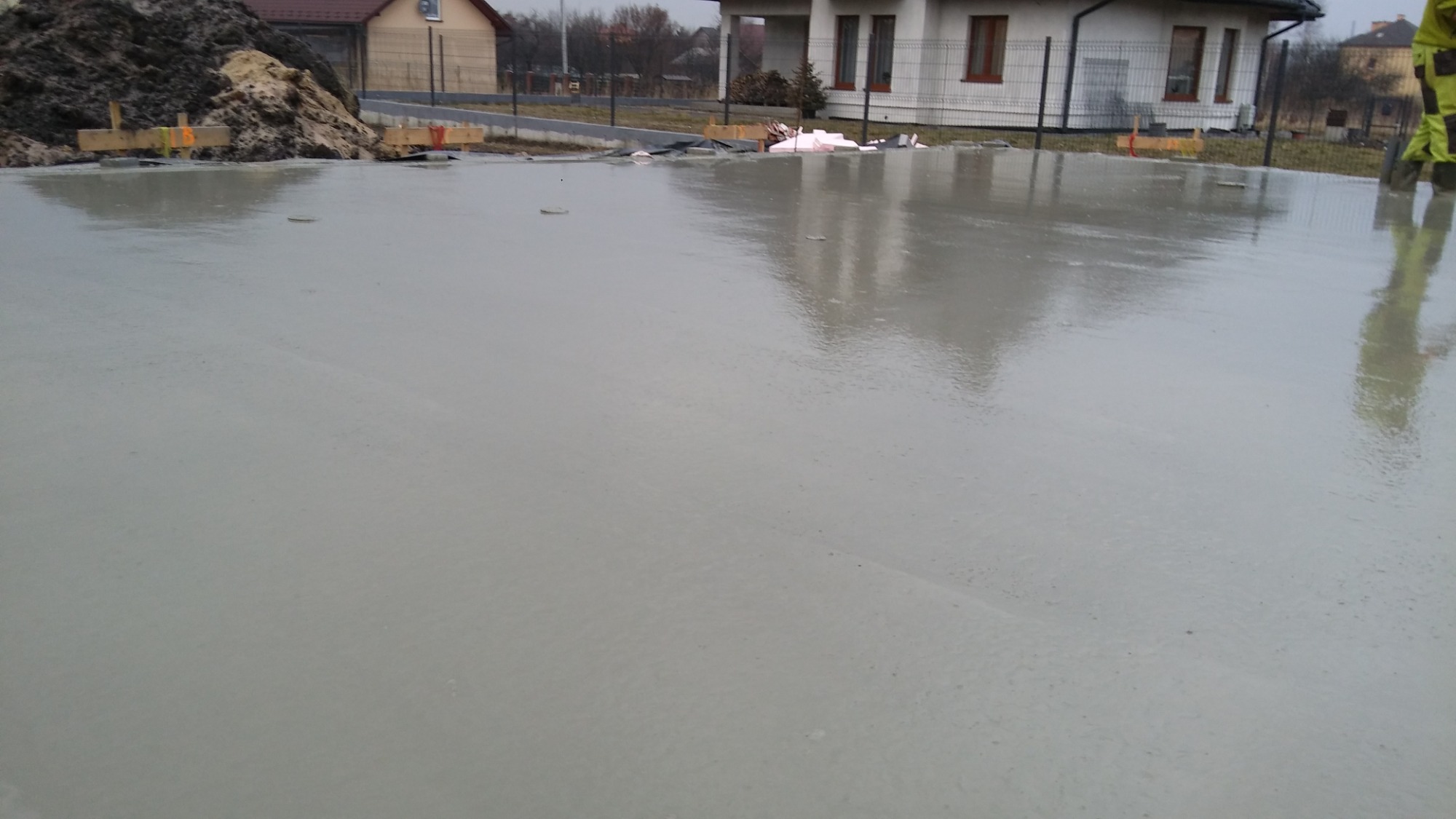 Świeżo wylany, mokry betonowy fundament, odbijający budynek w swojej gładkiej powierzchni, z widocznymi drewnianymi elementami szalunku i stertą ziemi w tle.