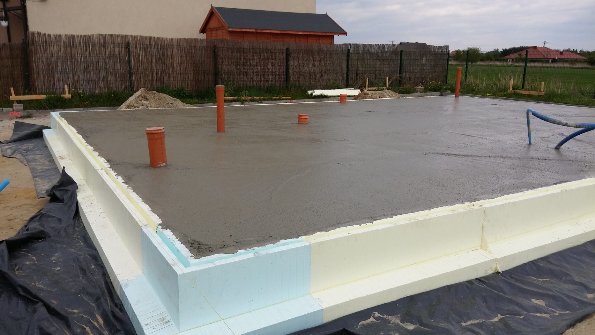 Wylany beton na przygotowany szalunek z izolacją termiczną, wystające rury kanalizacyjne, widoczny teren budowy z domkiem narzędziowym w tle.